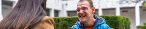 een man man met verstandelijke beperking in rolstoel tegenover een begeleider. De man lacht.
