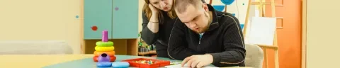 jongen met verstandelijke beperking aan een tafel. Hij doet een werkje. Naast hem staat een begeleister die meekijkt.